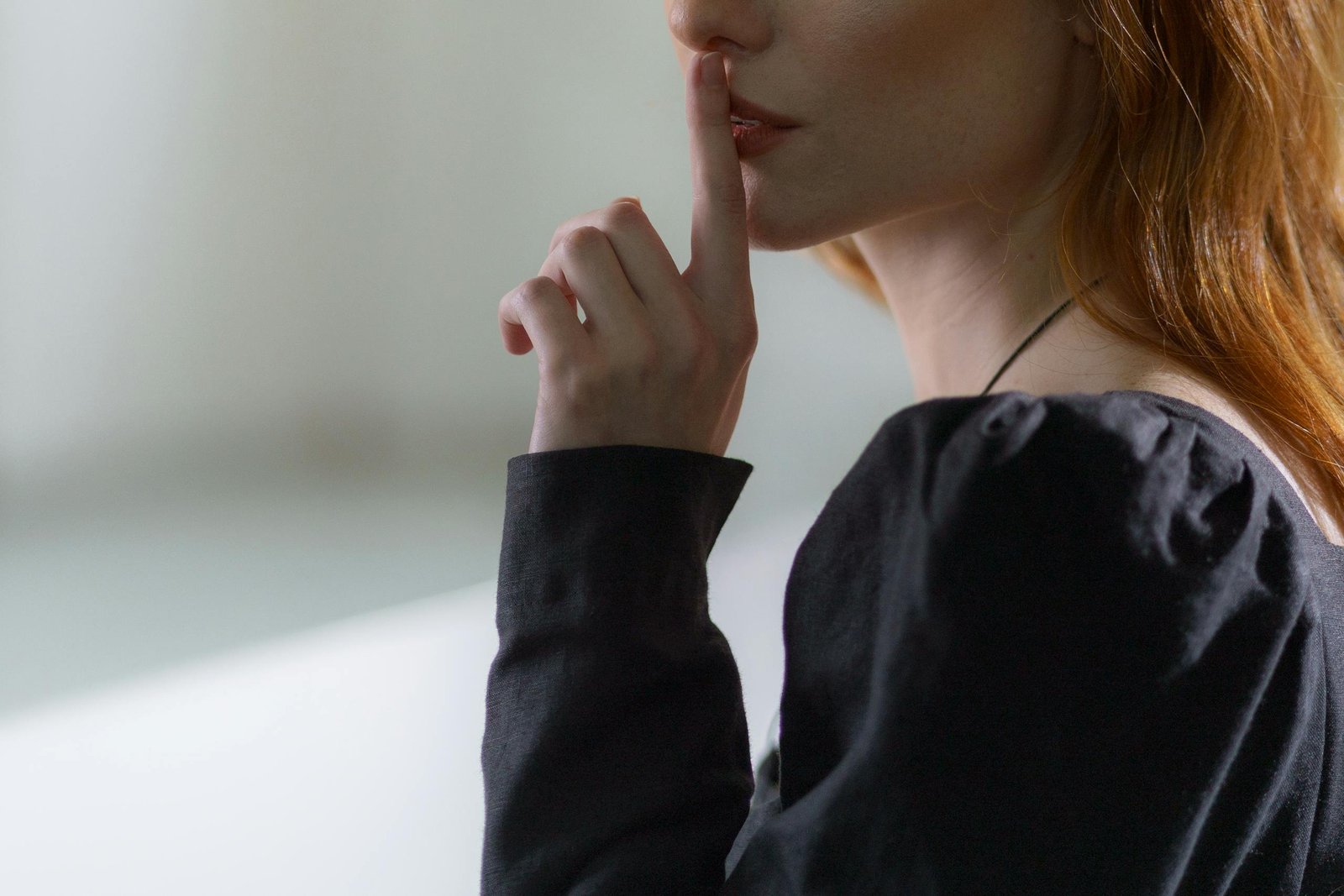 Close-up of woman suggesting silence with finger on lips, wearing a black top.