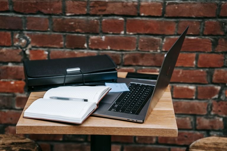 Open laptop and notebook on a wooden table by a brick wall, perfect for remote work inspiration.